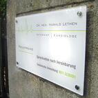 Weißes Praxisschild aus Acrylglas, montiert mit runden Edelstahl-Punkthaltern an einer Praxiswand in Mainz.